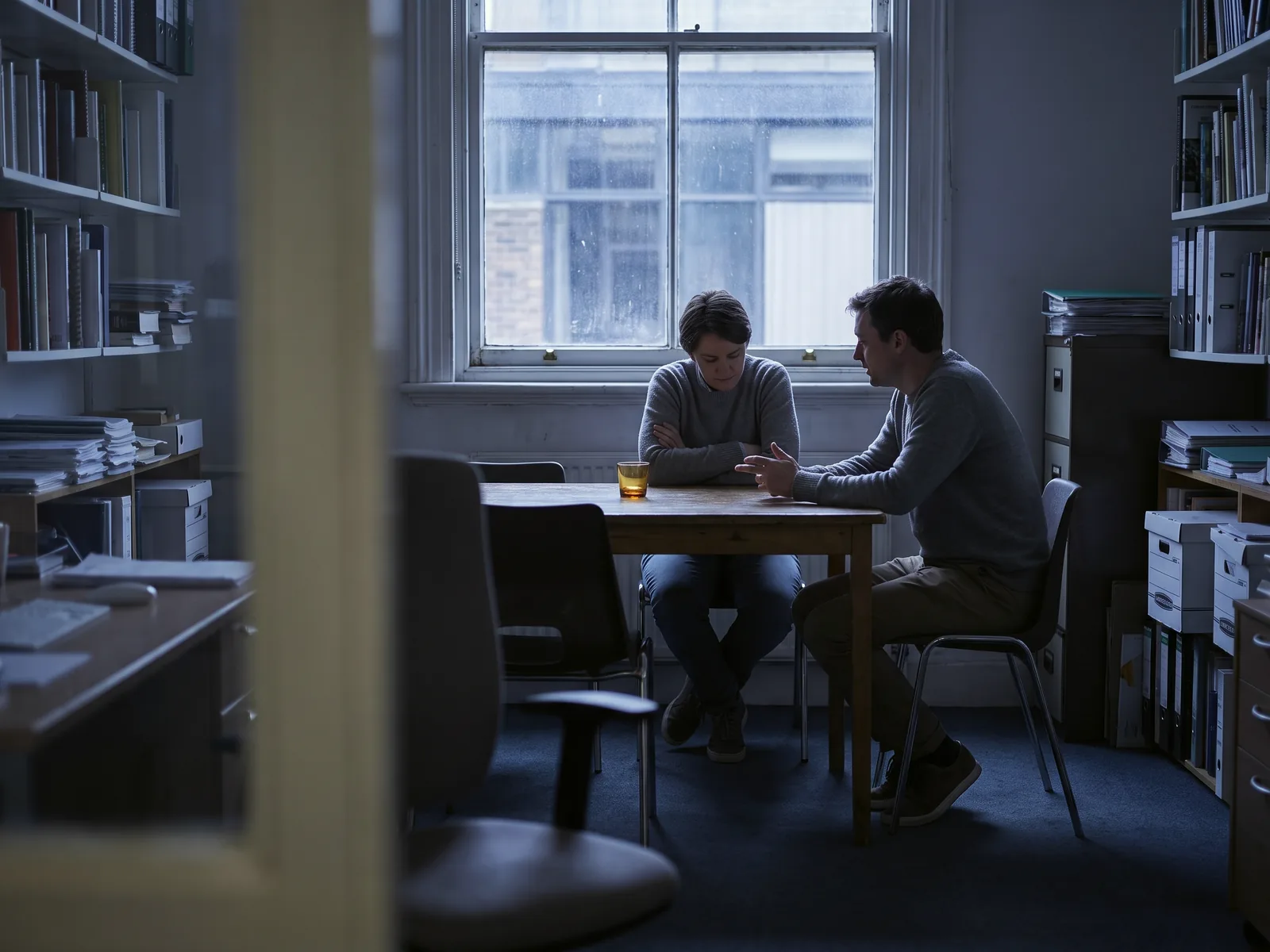 Two colleagues talking across a desk in a small office, seen from the doorway.