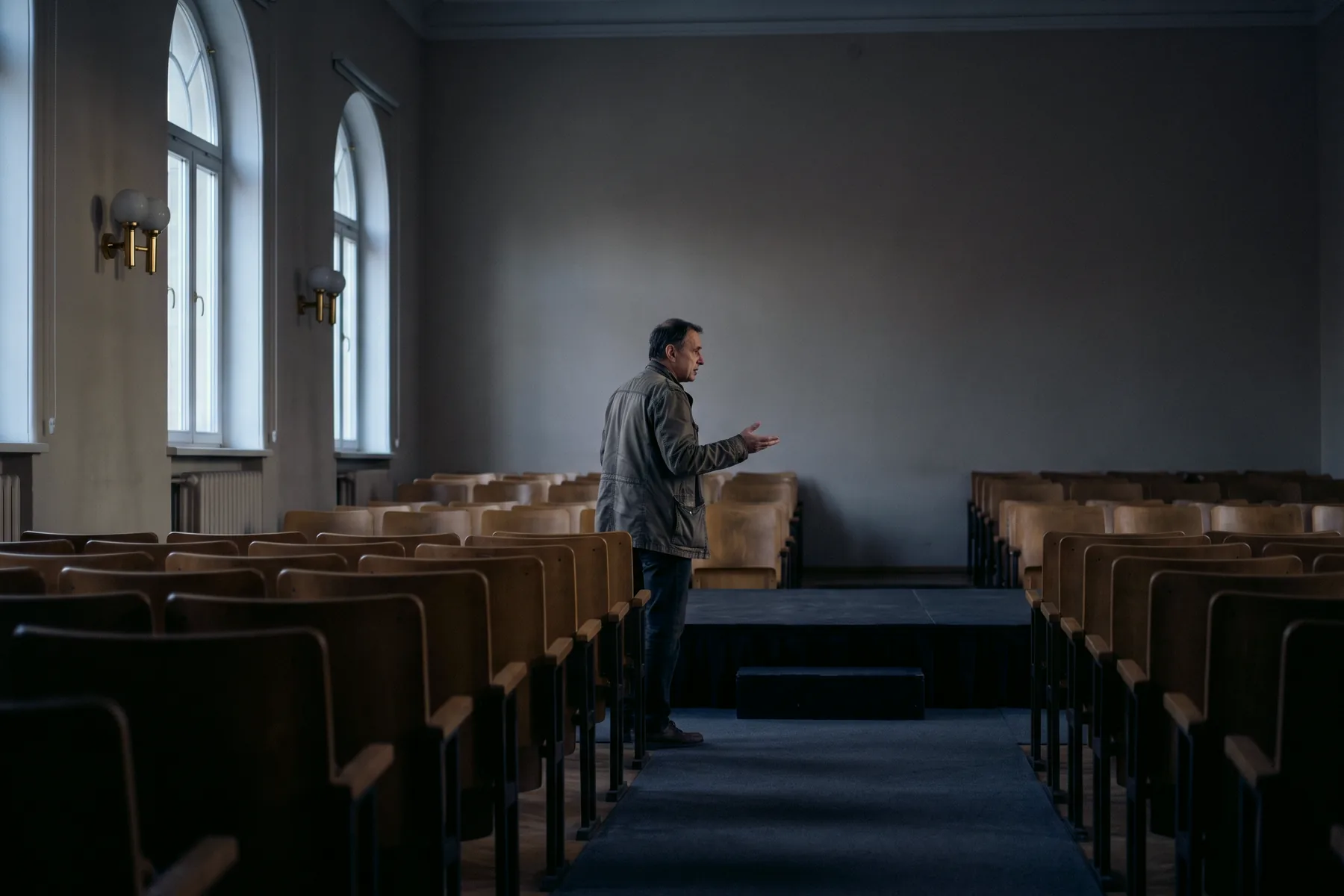 A person alone on a low stage in an empty lecture room, mid-sentence, rehearsing before the audience arrives.