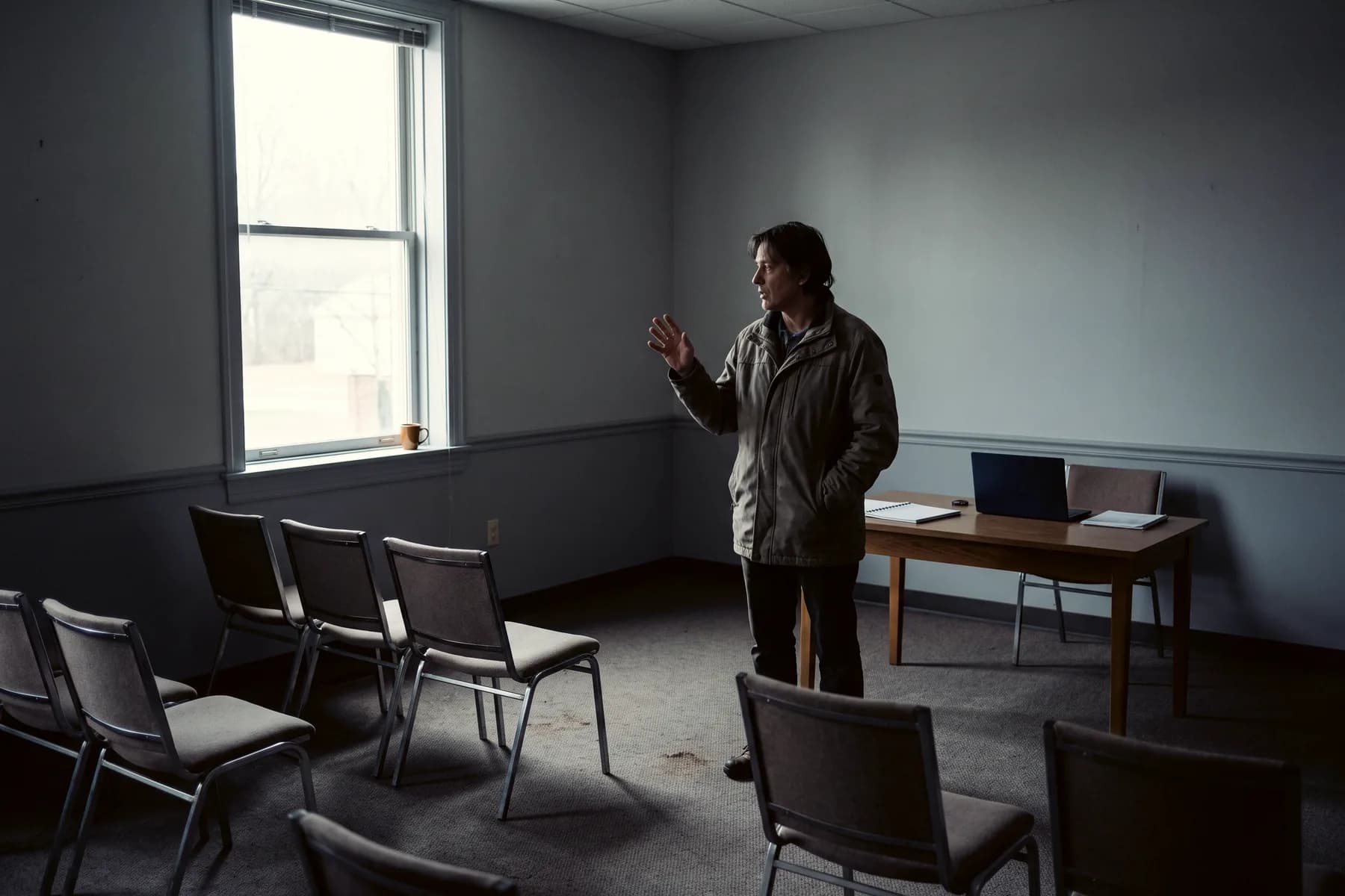 A person alone in a meeting room, rehearsing to empty chairs, one hand mid-gesture.